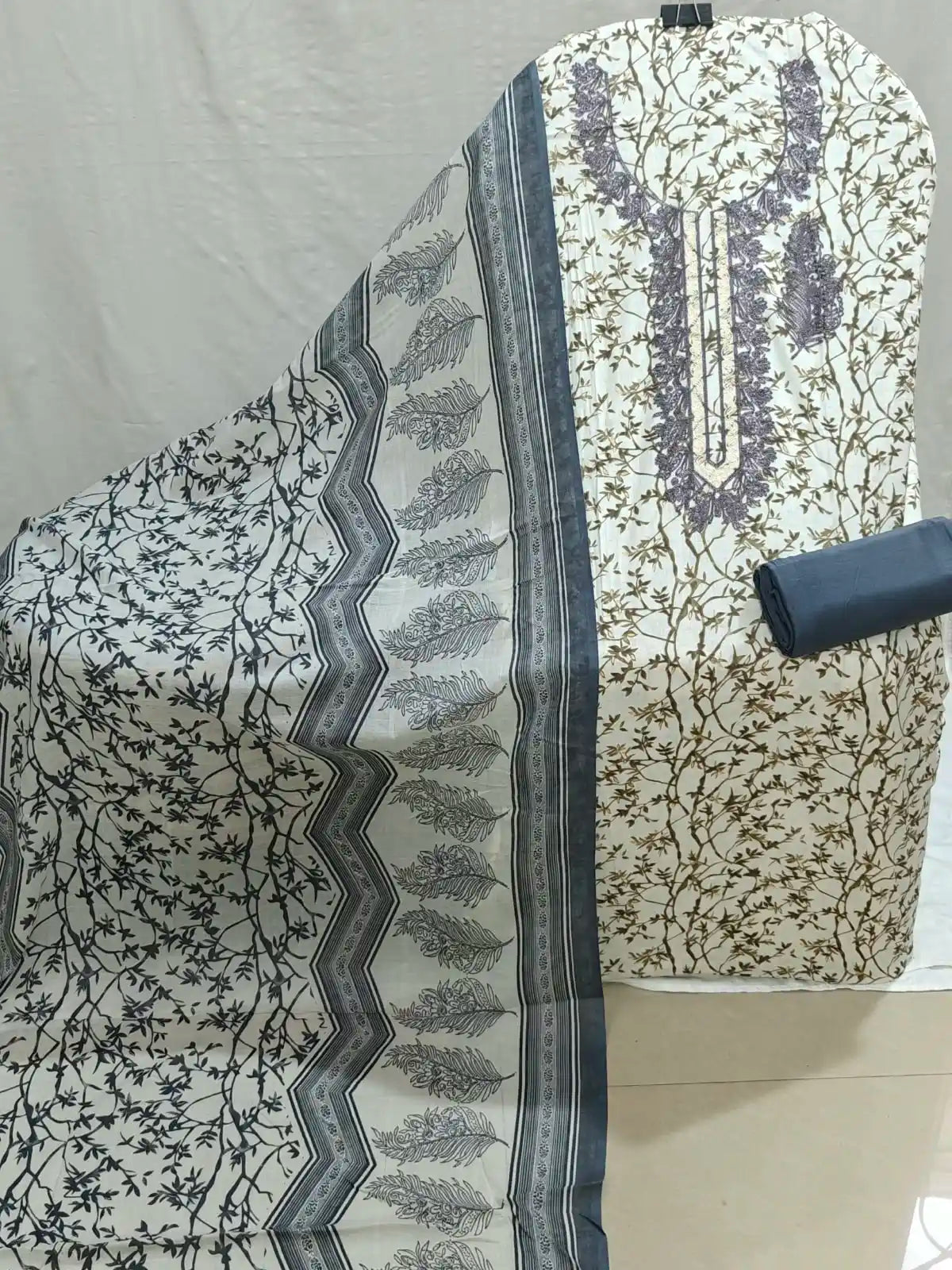 A folded and draped piece of printed fabric, likely a salwar suit material, featuring a cream base with black floral patterns, a gray and black zigzag border, and a purple printed neckline design on the reverse side.