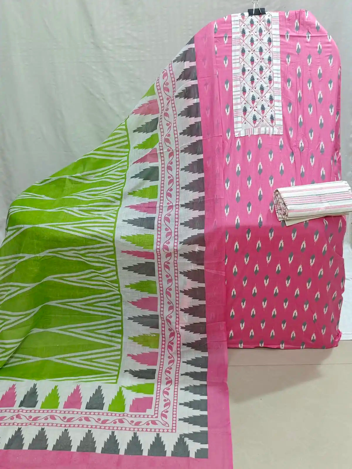 A pink cotton salwar suit material set with a printed kurta piece and a matching dupatta. The kurta features a pink body with small white and gray ikat motifs, a white embroidered yoke, and a patterned border.