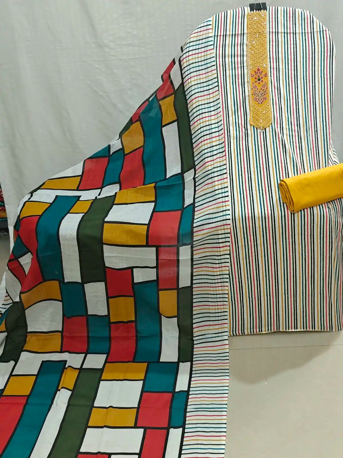 A multicolor printed and striped fabric set displayed on a chair, featuring a large draped dupatta with a geometric block pattern in red, yellow, teal, green, and white, alongside a matching striped kurta blouse with a yellow embroidered yoke.