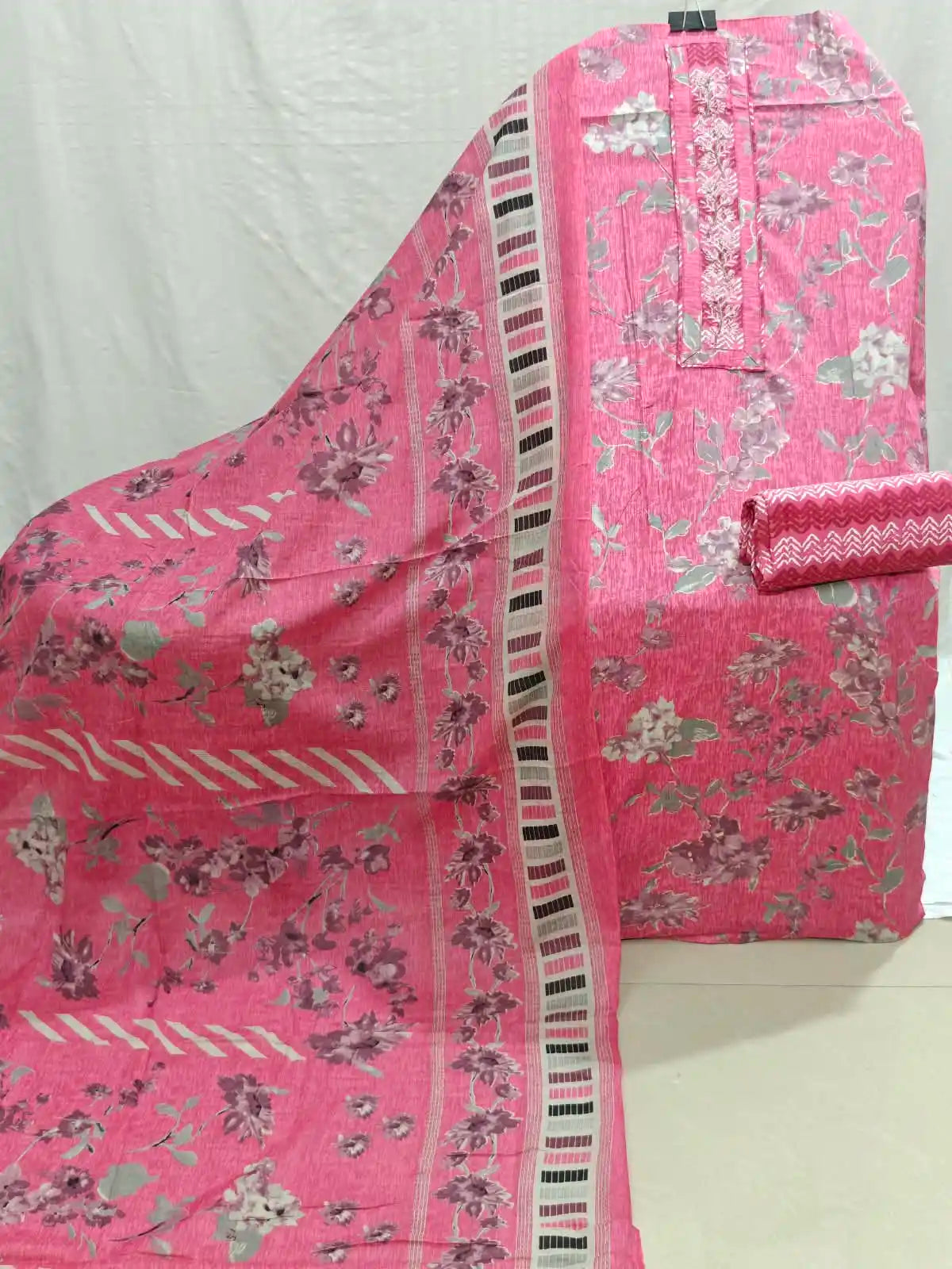 Pink floral printed fabric set with a matching dupatta draped over a folded garment piece, featuring a floral pattern and striped border details.