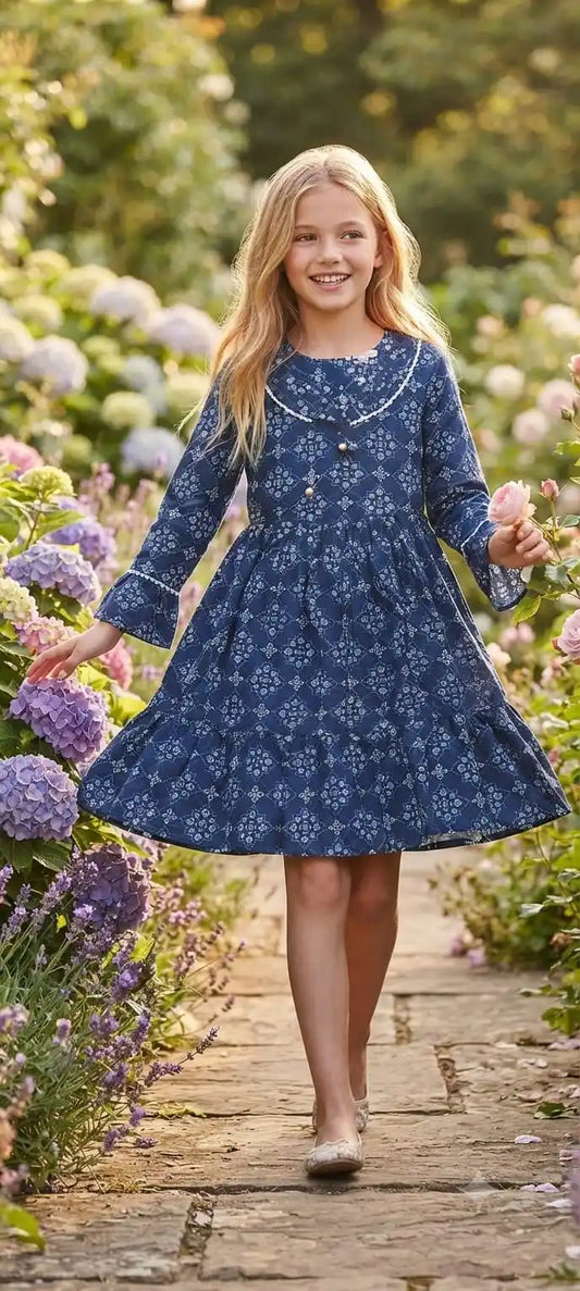 A blonde girl smiling and holding a pink rose while walking on a pathway, wearing a navy blue dress with fine white geometric tile patterns and lace-trimmed bell sleeves.