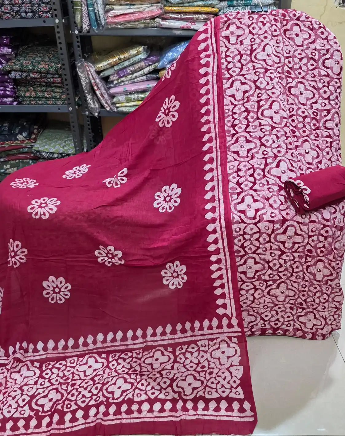 Unstitched magenta-red cotton suit material featuring an all-over white four-petal geometric batik print interspersed with subtle mirror-work details, paired with a solid magenta bottom fabric roll, and a matching draped dupatta adorned with white eight-petal floral motifs and a broad geometric batik border.