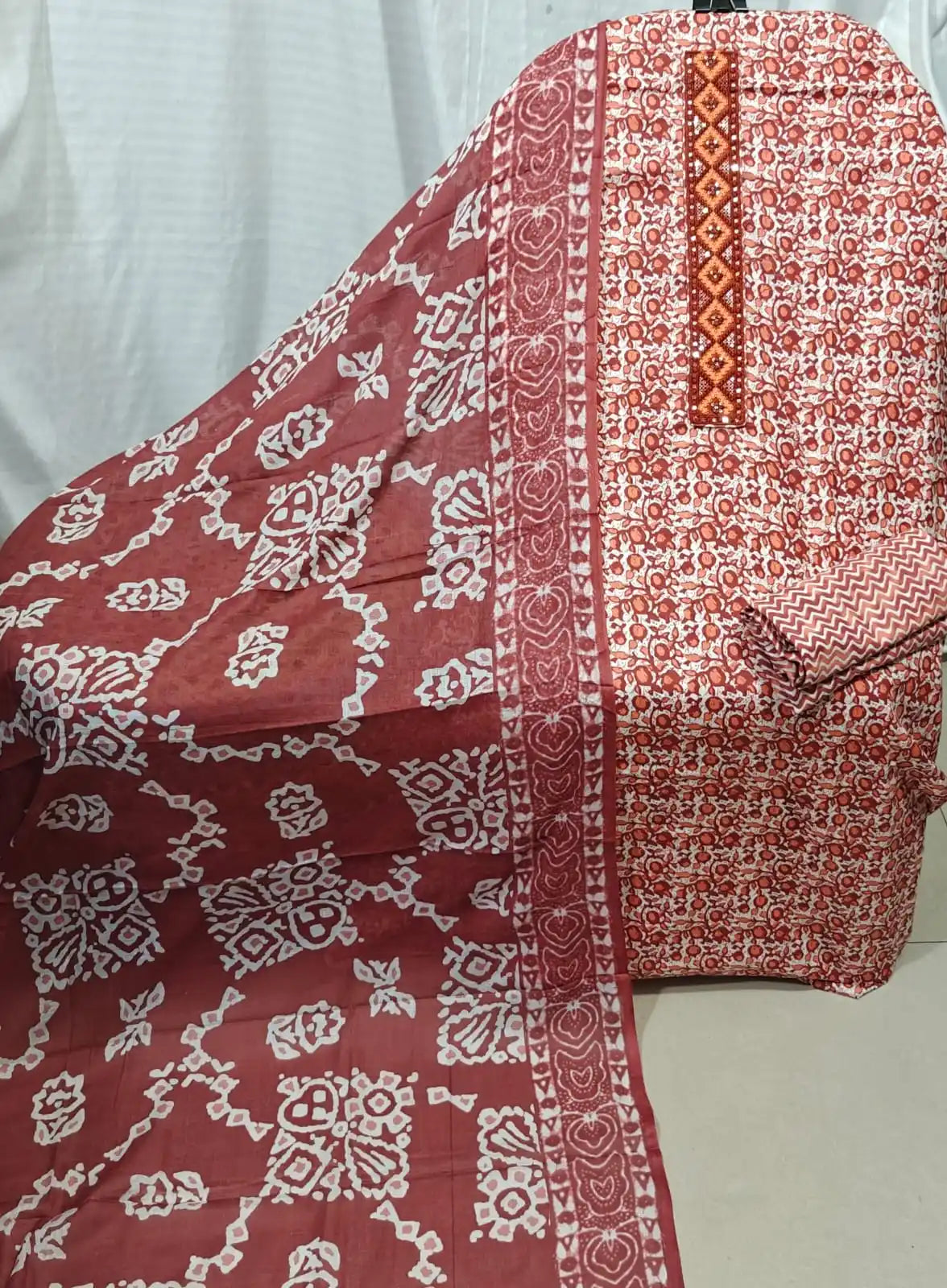 A rich rust red cotton unstitched dress material by ASC, showing an intricate micro floral print, a detailed orange and red embroidered neck panel, and a matching rust dupatta with white geometric batik art.