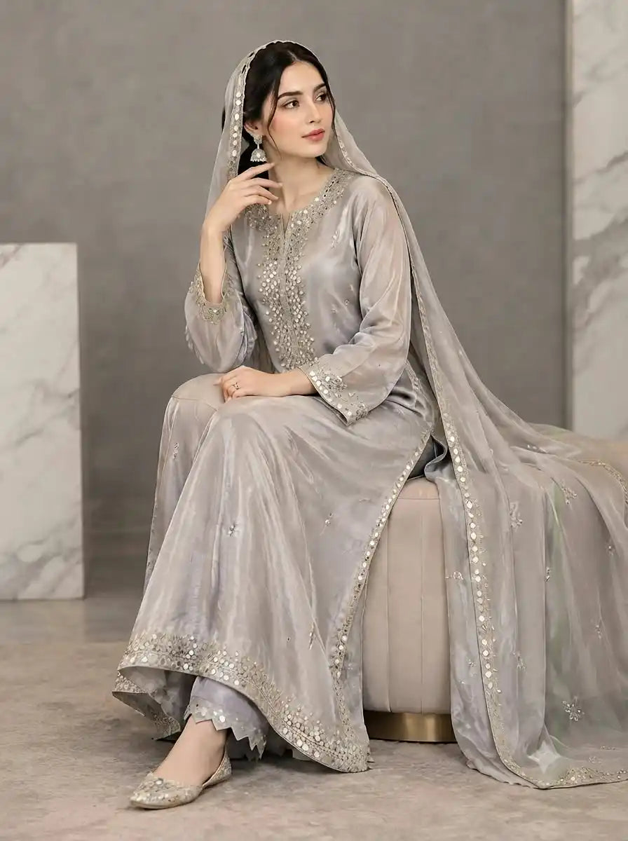 A woman seated on a cushion wearing a light silver-gray traditional outfit with ornate embroidery and a matching Duppatta