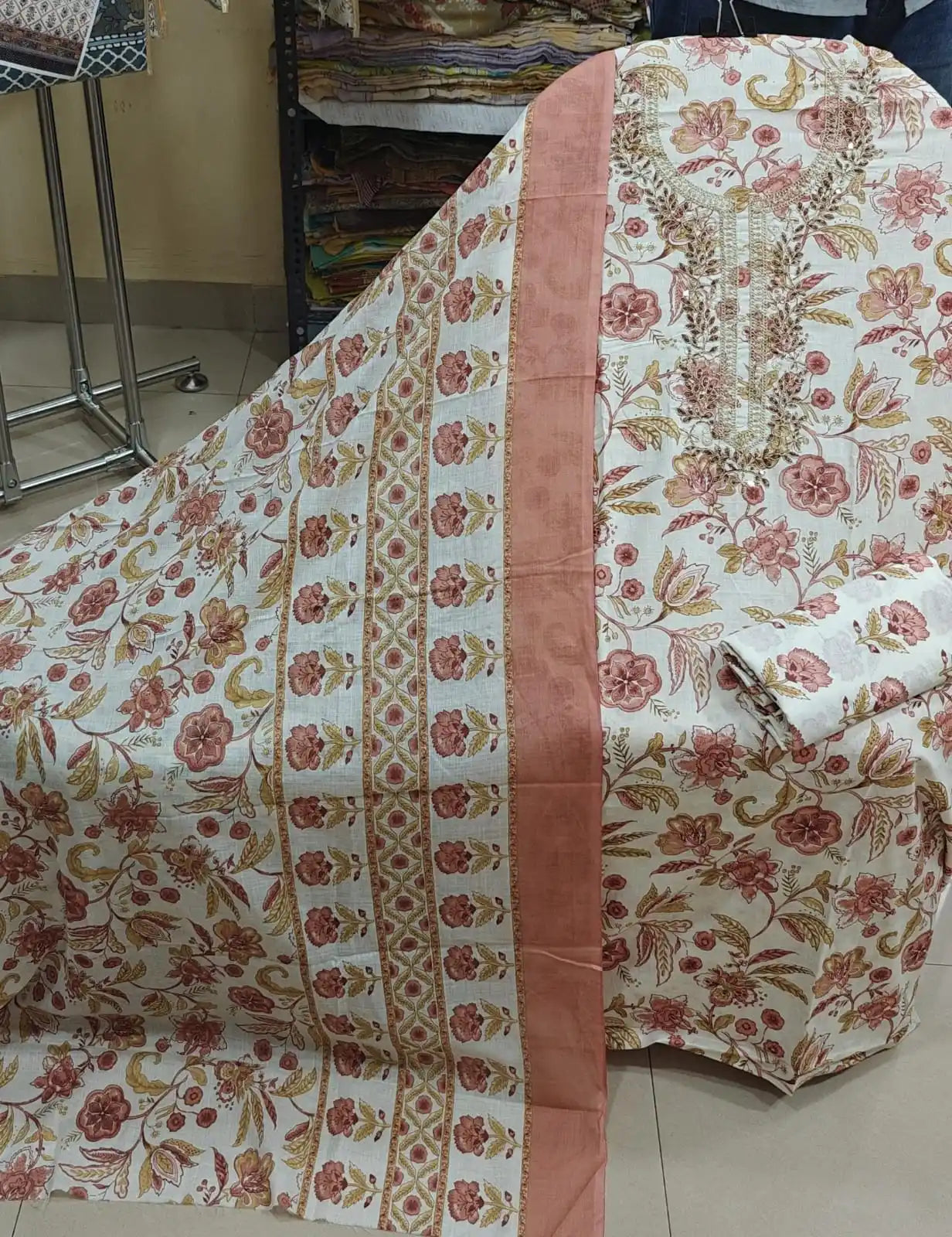 Cream white unstitched cotton suit displaying peach-pink floral patterns and an intricate embroidered neckline.