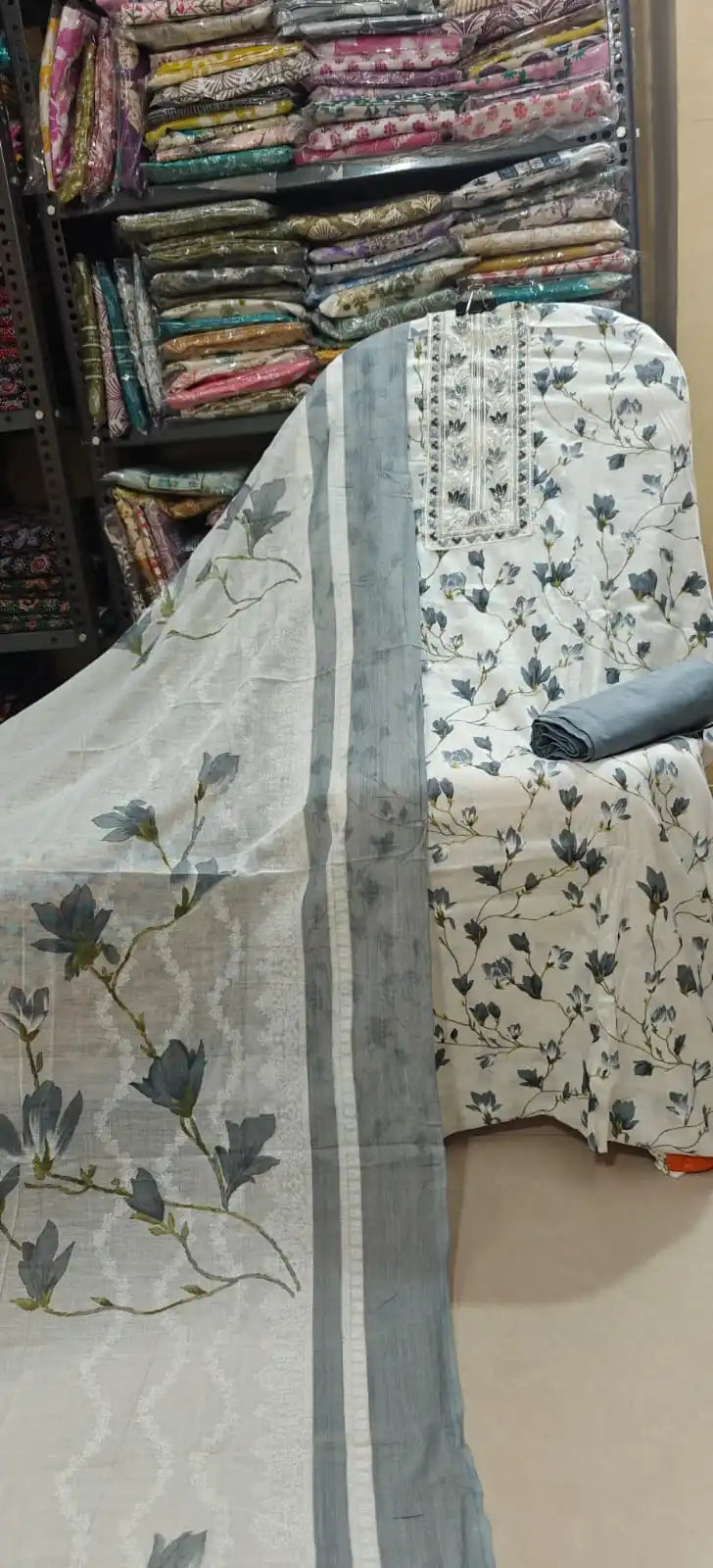 Premium white cotton dress material showcasing a dark gray floral print and an ornate embroidered neckline, accompanied by a solid gray bottom fabric and a matching printed dupatta.
