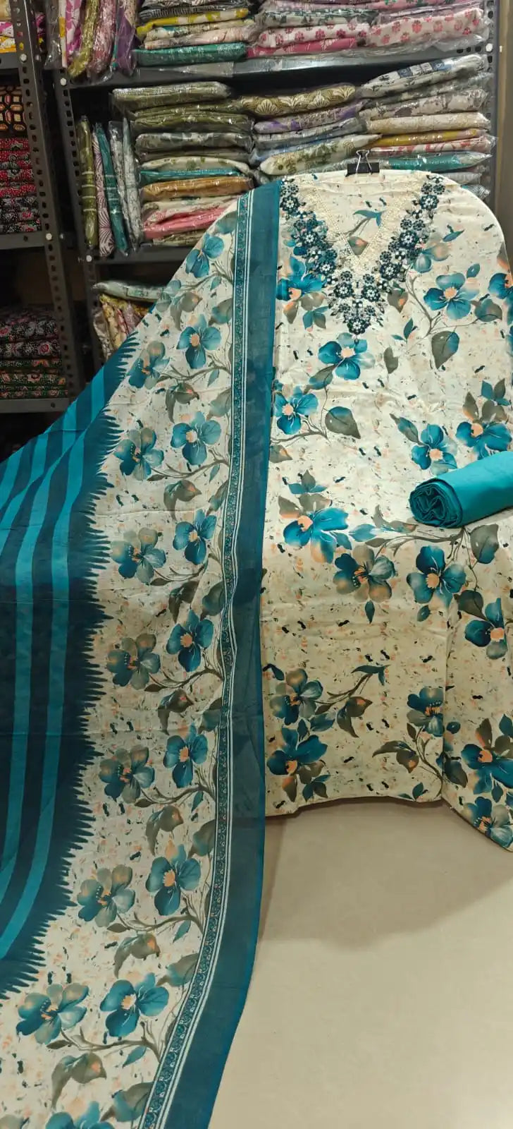 Cream cotton suit material featuring a striking turquoise blue floral print, enhanced by a heavy teal blue embroidered V-neckline design, paired with a solid teal bottom and a matching striped dupatta.