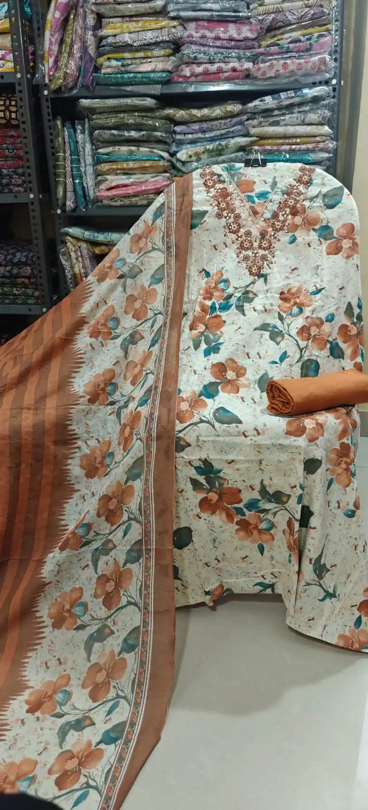 Cream-toned unstitched cotton suit embellished with a rich rust-orange floral print and an elaborate brown thread embroidered neckline, styled with a solid rust-colored bottom and a matching dupatta.