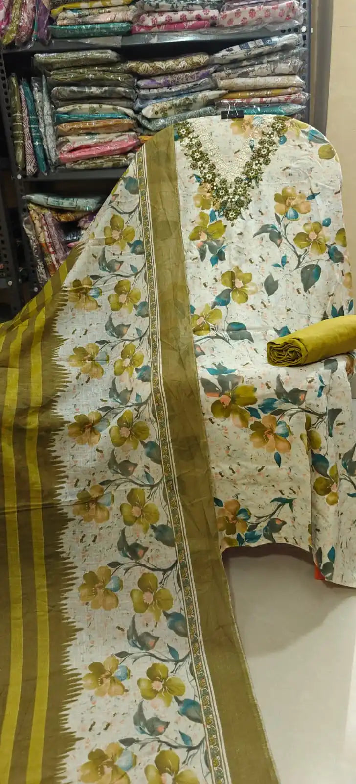 Cream unstitched cotton suit material with a prominent olive green and brown floral print, featuring a heavy V-shaped beaded or embroidered neckline, paired with a solid olive green bottom fabric and a matching printed dupatta.