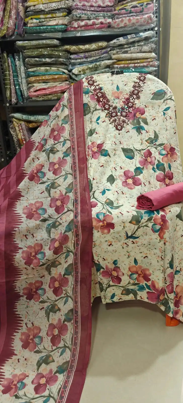 Off-white cotton dress material adorned with large maroon and purple floral motifs, styled with an intricately embroidered dark maroon neckline, a solid pinkish-maroon bottom roll, and a coordinated printed dupatta.