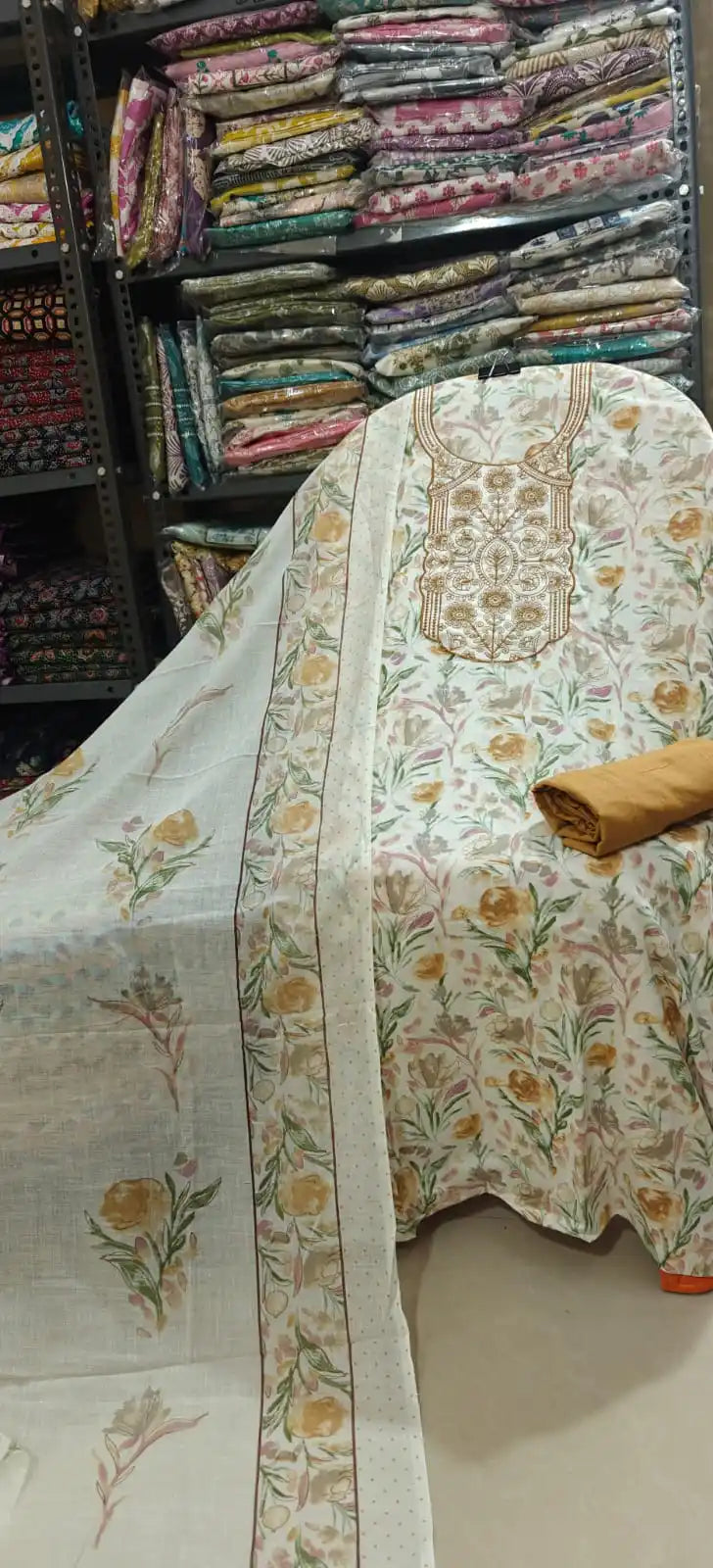 Premium white cotton unstitched suit featuring a subtle all-over beige floral vine print and a detailed round embroidered neck patch in gold-beige threads, complete with a solid beige bottom and a matching dupatta.