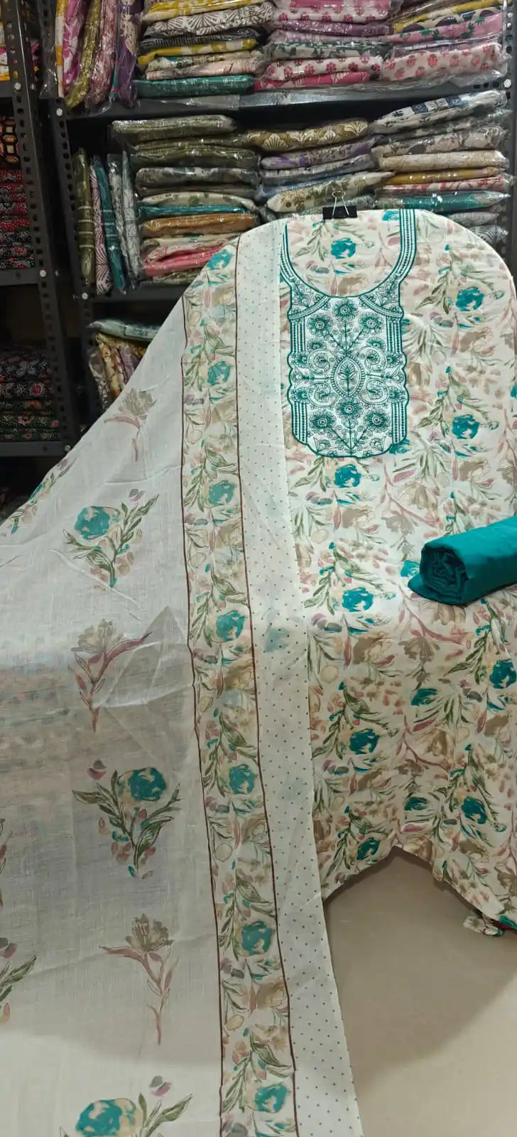 Elegant white cotton dress material showcasing soft teal and muted brown floral prints, highlighted by a rich teal green embroidered neckline panel, paired with a solid teal bottom fabric and a printed dupatta.