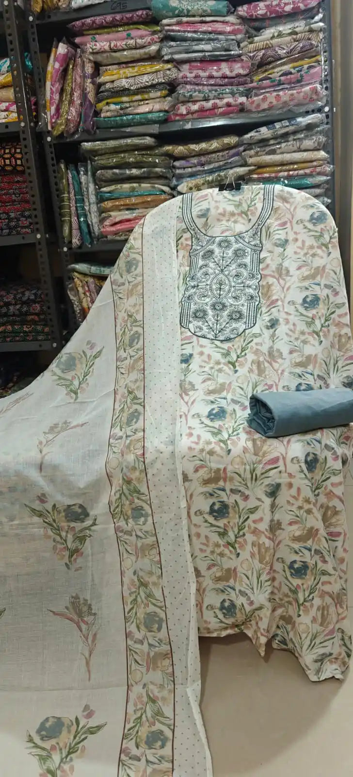 White unstitched cotton salwar kameez material with a delicate multi-colored floral pattern, featuring a sophisticated gray-green embroidered neck design, a solid slate gray bottom, and a matching dupatta.
