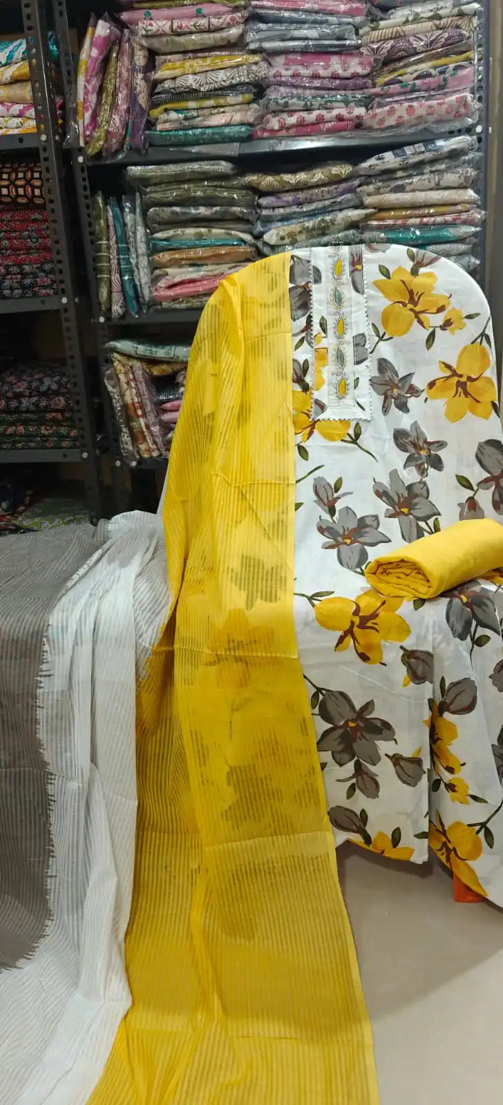White cotton dress material featuring large vibrant yellow and grey floral prints, styled with a simple embroidered neck border, a solid yellow bottom roll, and a dual-toned yellow and grey striped dupatta.