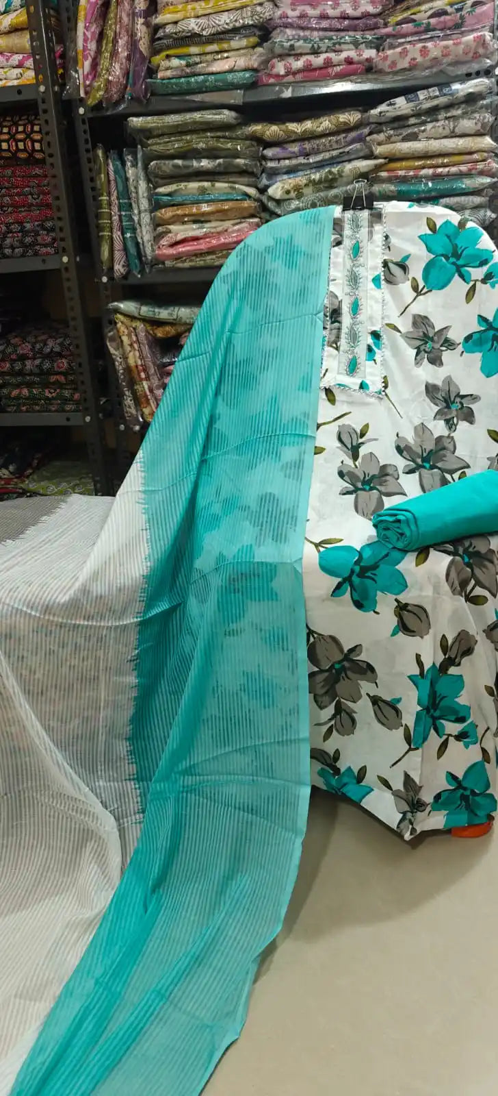White unstitched cotton suit material decorated with large cyan and grey floral prints, featuring a matching vertical embroidered neck patch with lace edges, paired with a solid cyan bottom fabric and a dual-toned cyan and grey striped dupatta.