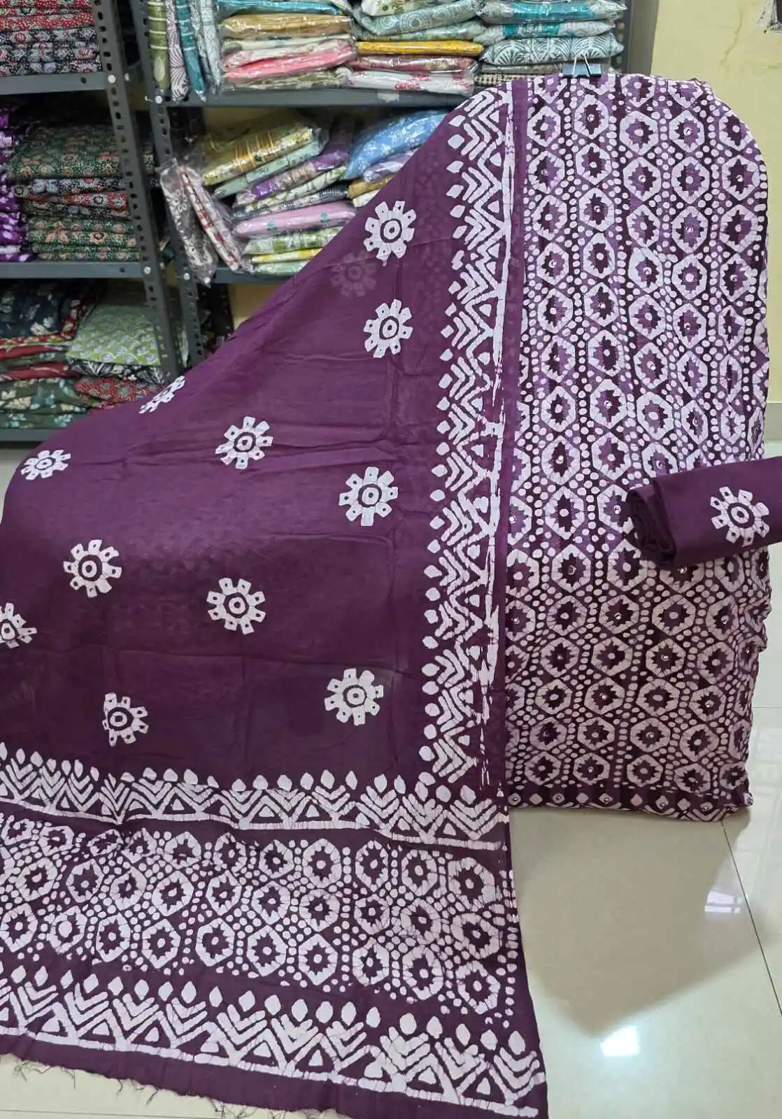Dark purple unstitched cotton dress material showcasing a dense white diamond-patterned batik print on the main shirt fabric, complete with a solid purple salwar piece, and a flowing purple dupatta decorated with white circular emblems and geometric edge patterns.