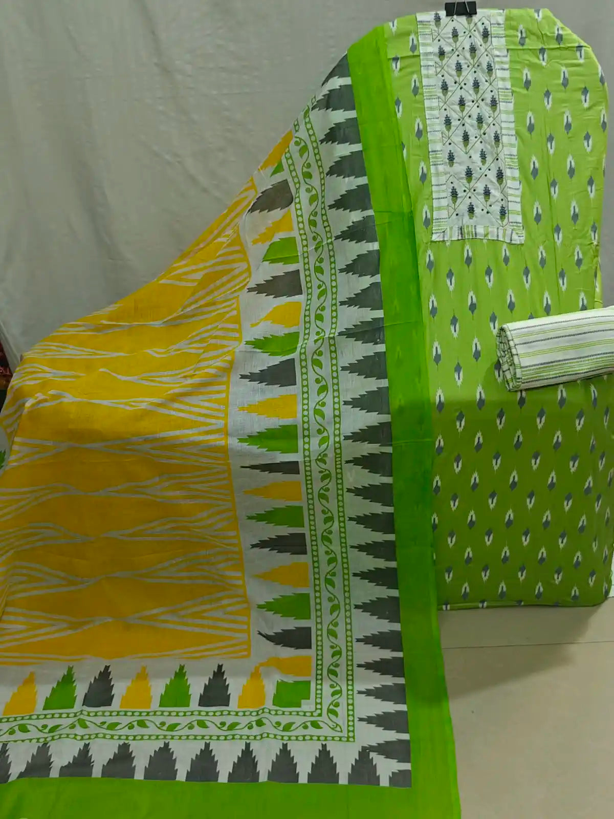 A folded and draped piece of bright green fabric with small white and blue motifs, shown alongside a matching patterned dupatta or scarf in yellow, green, gray, and white. A folded striped fabric piece is also visible on the right side, all arranged against a plain light backdrop.