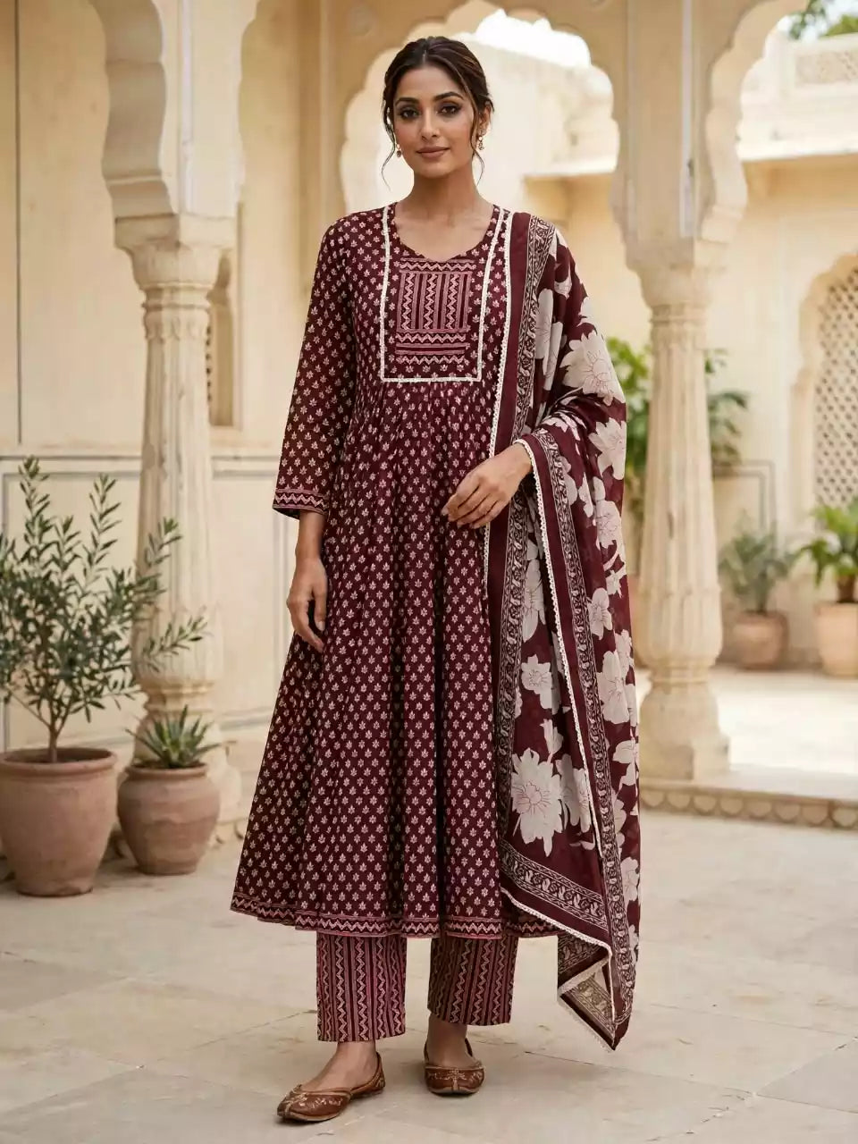 A woman stands in a courtyard wearing a traditional Indian outfit consisting of a maroon kurta with white floral prints and embroidery, matching printed pants, and a large printed dupatta.