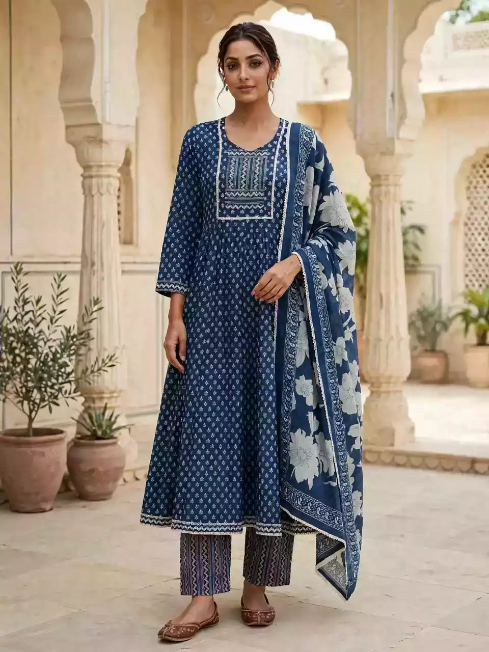 A woman stands in a courtyard wearing a navy blue printed salwar kameez suit with white border embroidery, matching patterned trousers, and a navy dupatta with white floral prints.
