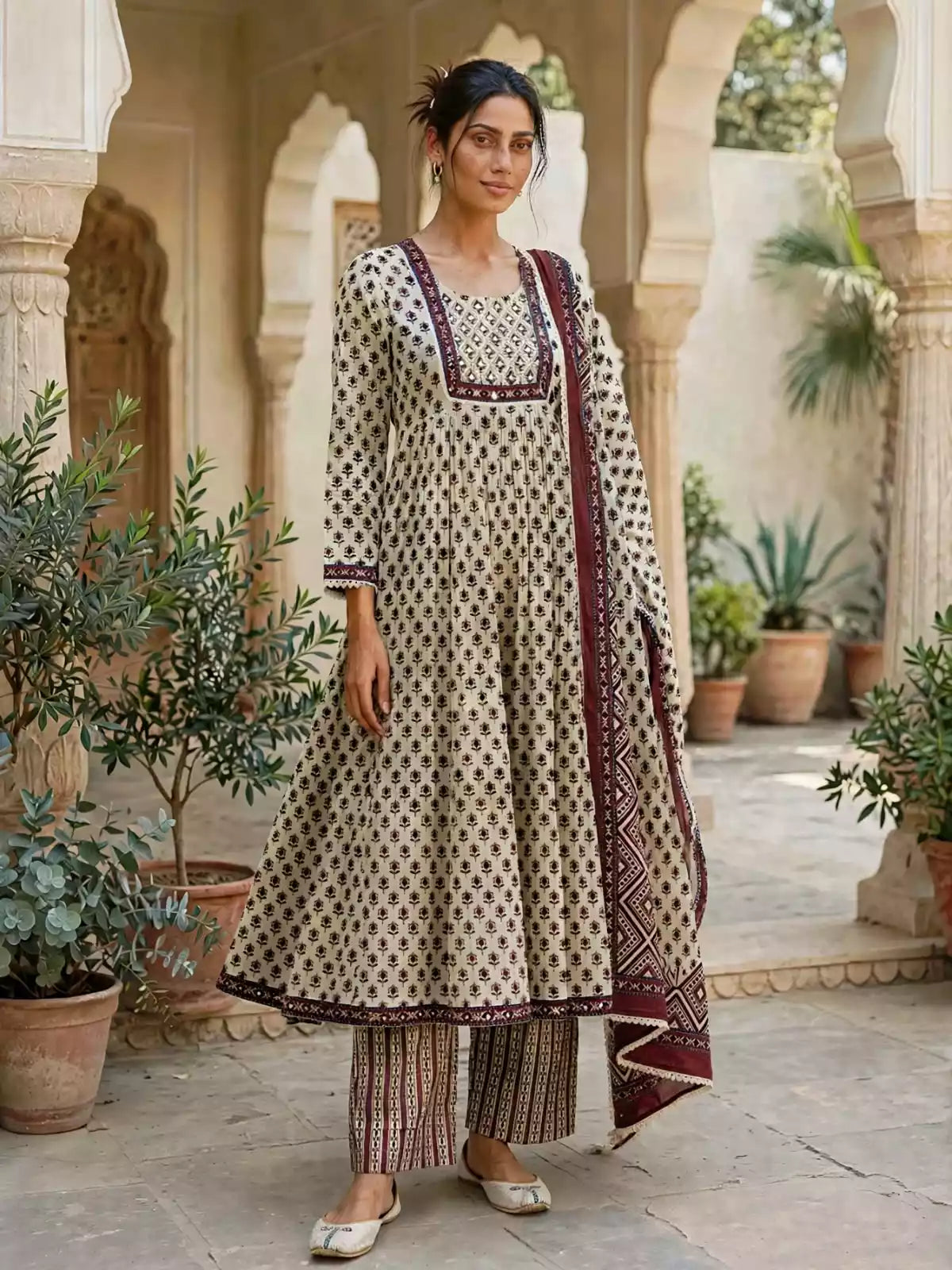 A woman models a traditional Indian salwar kameez suit in an outdoor courtyard with arches and potted plants. The outfit consists of a beige kurta with black block prints and a maroon border, matching striped pants, and a maroon patterned dupatta.