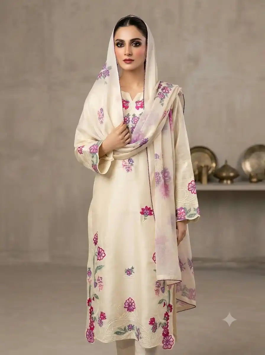 A woman modeling a cream-colored traditional outfit with pink and green floral embroidery, featuring a matching dupatta, set against a neutral gray studio background.
